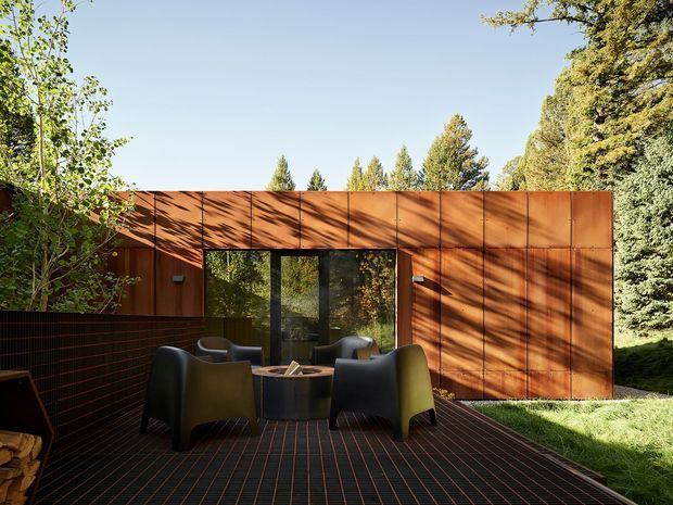 A modern single-storey outdoor terrace or courtyard framed by a long rust-coloured metal-clad wall. The terrace has dark tiled or grid-patterned flooring, a low dark privacy wall, four sculptural lounge chairs, and a central round fire pit or table. A large glazed opening connects the terrace to the wooded garden, with mature trees and strong dappled sunlight creating a secluded setting.