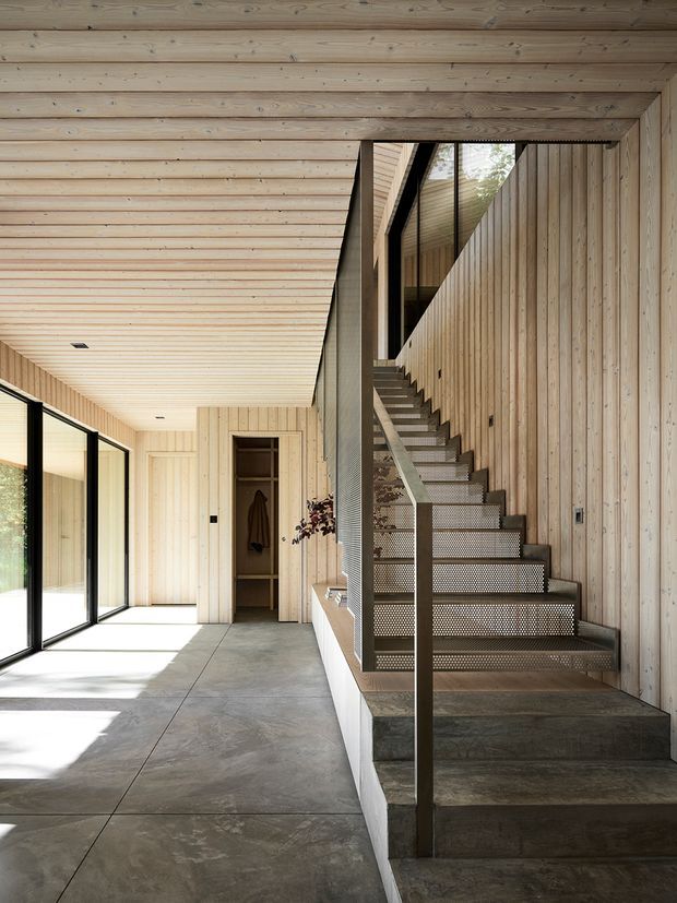 A bright, minimalist interior hallway and stairway clad extensively in pale vertical and horizontal timber boards. A dark metal-framed open staircase with perforated metal risers rises beside a tall glazed void, while full-height black-framed glazing lines the left side. The floor is a smooth grey concrete finish, and a small built-in storage area is visible beneath the timber wall finish. Strong natural light creates an airy, warm contemporary atmosphere.