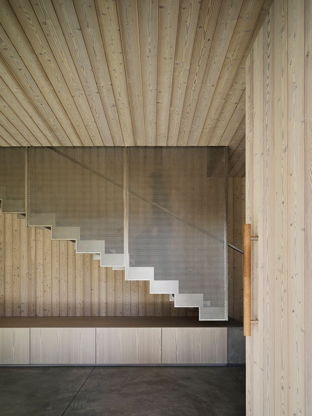 Interior architectural photograph dominated by a sculptural straight-flight staircase with perforated metal balustrades and pale timber-clad walls and ceiling. The stair descends toward a dark concrete floor, with minimal built-in cabinetry beneath and a simple timber handrail. The composition highlights the junction of timber lining, metal detailing, and contemporary joinery.