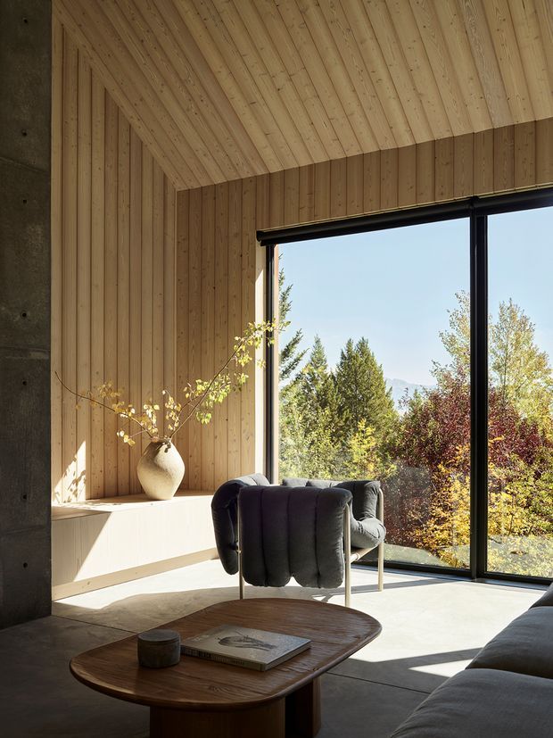 A warm, minimalist living room with extensive vertical timber wall panelling and a matching sloped timber ceiling. A large black-framed glazed opening overlooks trees and distant mountains, while a built-in timber window seat, dark sculptural lounge chairs, a rounded wooden coffee table, and a partial sofa create a refined, nature-connected interior. A concrete structural wall is visible at the left.
