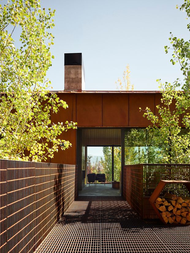A modern single-storey building viewed from a raised outdoor walkway or terrace. The facade is clad in weathered rust-brown metal panels, with a dark metal chimney and a large glazed opening leading into an interior sitting area. A metal grating floor, slatted metal side screens, timber railings, and a stacked firewood storage feature create a strong industrial, earthy character. Leafy trees frame the structure and sunlight casts patterned shadows across the walkway.