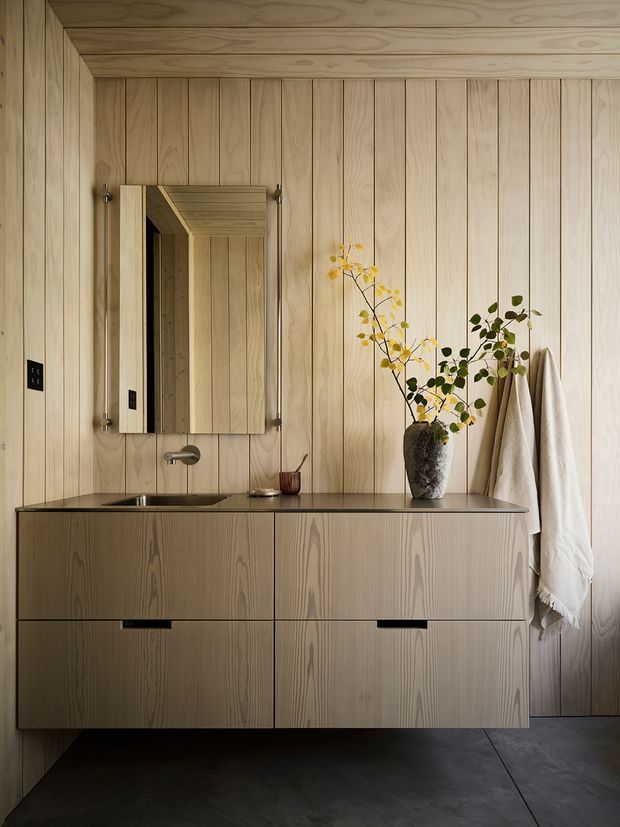 A compact, refined bathroom vanity area clad in pale vertical timber panelling across the walls and ceiling. A wall-mounted timber vanity with integrated dark stone benchtop, inset basin and minimalist tap sits beneath a rectangular mirrored cabinet. Natural branches in a textured vessel and draped towels add organic softness, while dark flooring and black hardware provide contrast.