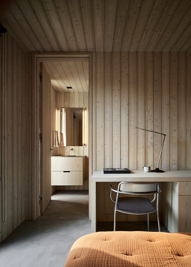 A warm, compact timber-lined bedroom with a built-in desk and perforated metal chair in the foreground. The room features vertical light-wood wall panelling and a matching plank ceiling, a smooth grey concrete floor, minimalist desk accessories, and a partially visible bed with a rust-orange quilt. Through the doorway is a small vanity area with a mirror, basin, and pale built-in cabinetry.