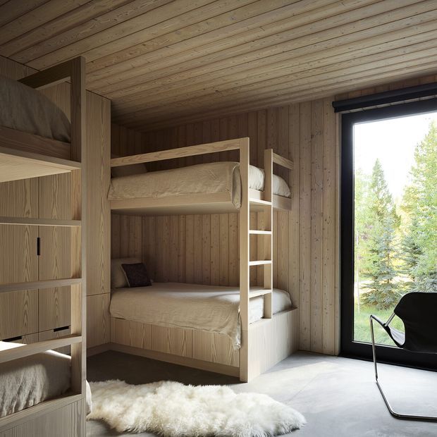 Compact bunk bedroom with custom built-in timber bunk beds and shelving, full-height vertical timber wall and ceiling panelling, a large black-framed window, pale flooring, and a light sheepskin rug. The room has a warm, minimalist cabin-like character and overlooks trees.
