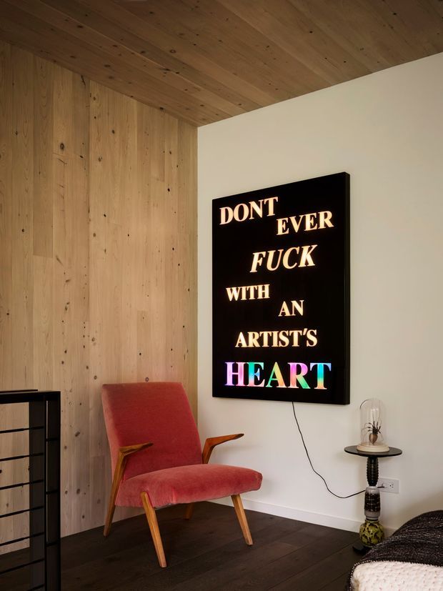 A cozy, compact interior corner featuring a red upholstered mid-century lounge chair with timber arms and legs, set against a vertical timber-panelled wall and warm timber ceiling. A large black illuminated typographic artwork hangs on the white wall, beside a small side table and lamp. Dark timber flooring and part of a bed are visible, suggesting a bedroom or bedroom-adjacent sitting area.