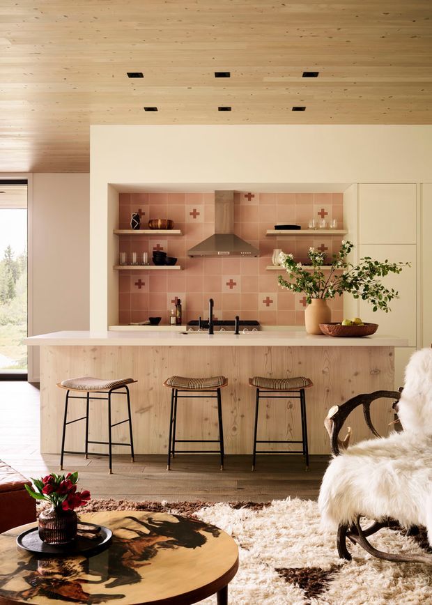 Warm, inviting open-plan kitchen and living area with a pale timber island and matching timber ceiling, pink tiled splashback with decorative cross motifs, stainless-steel rangehood, open shelving, and light cabinetry. Three black-framed stools line the island, while a sheepskin-covered rocking chair, textured rug, and round timber coffee table create a cosy, rustic-modern atmosphere. Natural light enters through large glazing at the left.