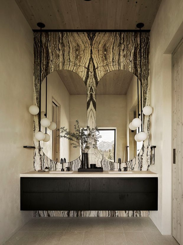 A refined double-vanity bathroom with a floating dark timber wall-mounted cabinet, pale stone benchtop, twin rounded-arch mirrors, and globe pendant lighting. Dramatic vertically veined black, cream and grey stone forms the feature wall behind the vanity. A timber-lined ceiling, pale plaster walls, tiled floor, and a mountain view through the rear window complete the calm, luxurious interior.