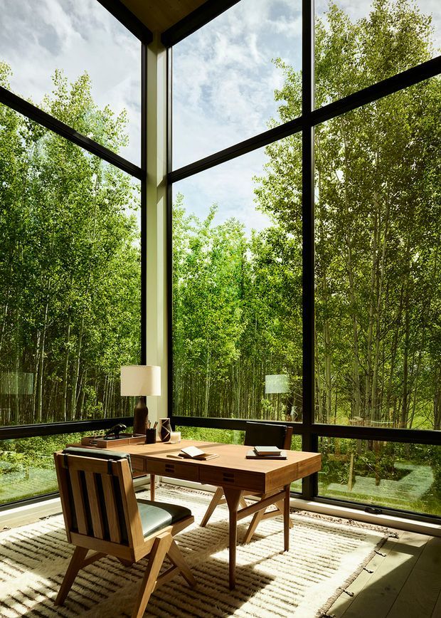 A compact home office positioned beside dramatic floor-to-ceiling corner glazing, with a timber desk, matching timber chair, desk lamp, books and stationery on a textured neutral rug. Black-framed window mullions create a strong grid, while dense green woodland is visible outside. The room has abundant natural light and a warm, nature-connected atmosphere.