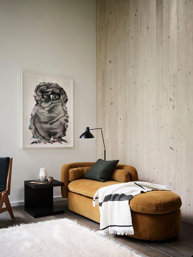 A warmly styled living room featuring a deep mustard-brown upholstered sofa with chaise, a black side table, a black adjustable floor lamp, a textured dark cushion, and a black-and-white throw. The room has a pale neutral painted wall, a large vertical light timber wall panel feature, dark timber flooring, a white shag rug, and framed owl artwork. The overall aesthetic is contemporary with tactile, inviting furnishings.