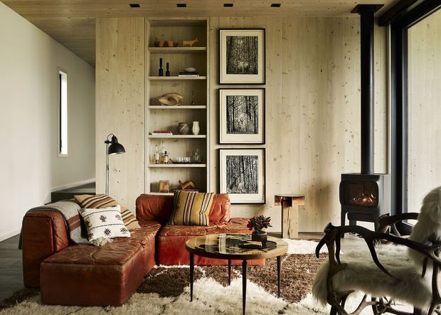A warm, compact living room with a deep brown leather sectional sofa, patterned cushions, a round coffee table, built-in timber shelving, pale timber wall and ceiling lining, and a black freestanding wood-burning stove. Three framed forest prints form a vertical feature beside the shelving, while a large black-framed window and a fur-covered lounge chair add contrast. The room has a rustic, cozy cabin-like atmosphere with strong natural materials and layered textiles.