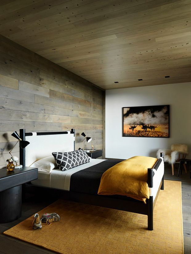 A warm, moody bedroom with a low black bed, white bedding, a black throw, and a mustard-yellow blanket. The room features extensive pale reclaimed timber wall and ceiling lining, a dark built-in bedside unit, black-and-brass articulated wall lamps, a large textured ochre rug, and a framed wildlife photograph. A lounge chair is visible in the corner.