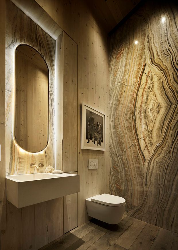 A compact, richly detailed bathroom featuring a floating rectangular vanity with integrated basin, an illuminated rounded-rectangle mirror, wall-mounted toilet, warm timber wall and ceiling panelling, timber flooring, and a dramatic vertically veined natural-stone feature wall and splashback. Recessed lighting creates a warm, intimate atmosphere.