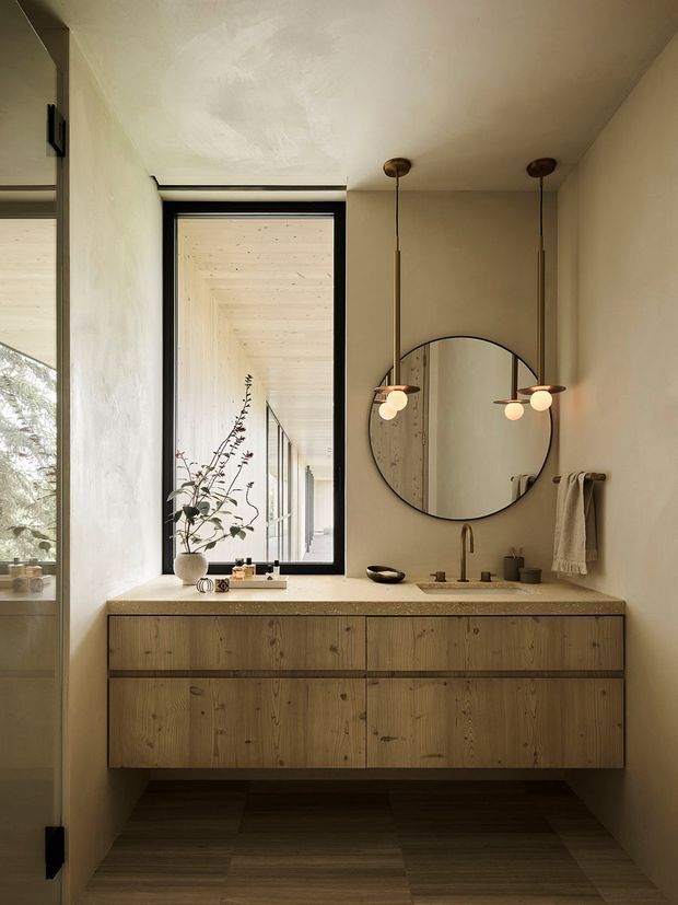 Warm, refined bathroom vanity area with a floating timber cabinet, pale stone benchtop, round black-framed mirror, brass-toned wall-mounted tapware, and twin pendant lights. A black-framed window opens to a light timber-lined corridor or exterior passage, while a textured towel and minimal accessories add softness. The room has plastered walls, a dark timber-look floor, and a calm natural-material palette.