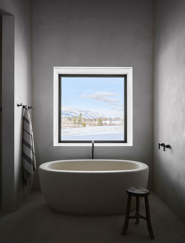 A serene, minimalist bathroom with a freestanding oval bathtub positioned beneath a square window. The room has smooth grey plaster walls, dark grey flooring, a white-trimmed black-framed window, a simple wall-mounted tap, and a small dark wooden stool. Snow-covered mountains and a winter landscape are visible outside.