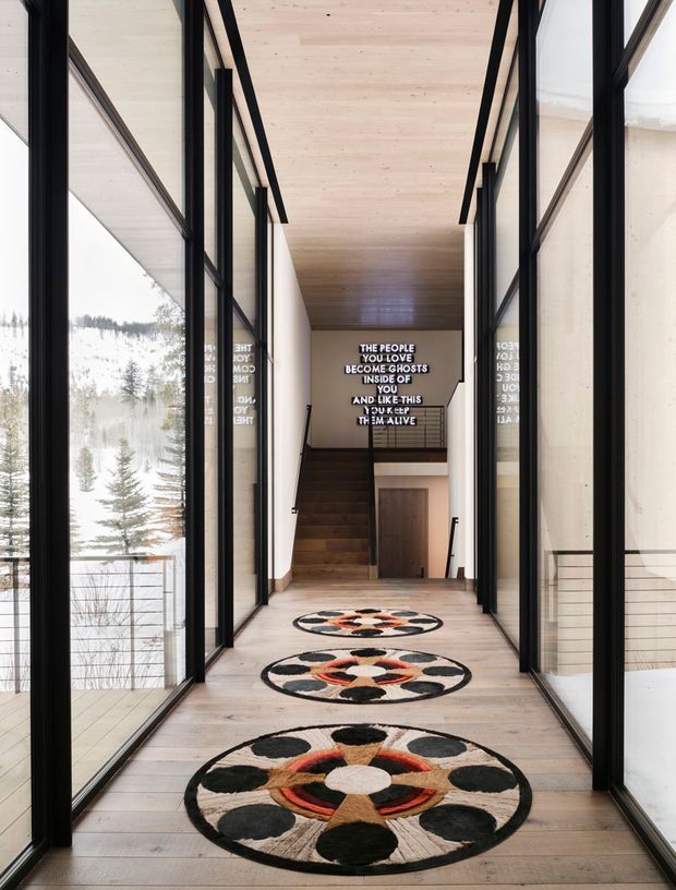 A long, narrow interior passage or landing with full-height black-framed glazing on both sides, overlooking a snowy wooded landscape. The corridor has pale timber flooring and a matching pale timber ceiling, creating a bright, minimalist architectural setting. Three circular patterned rugs lead toward a timber staircase and lower-level doorway. A bold illuminated typographic artwork is mounted on the far wall, with reflections visible in the glazing.