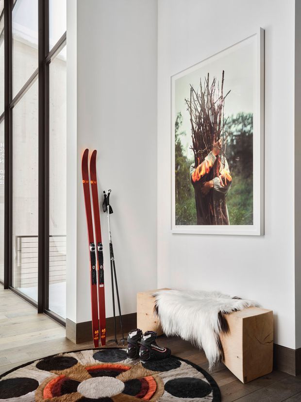A bright, tall modern entryway or hallway with floor-to-ceiling dark-framed glazing, white walls, light timber flooring, and dark trim. Skis and poles lean against the wall beside a sculptural timber bench draped with white fur, while a large framed photograph adds visual interest. A bold circular patterned rug anchors the foreground.