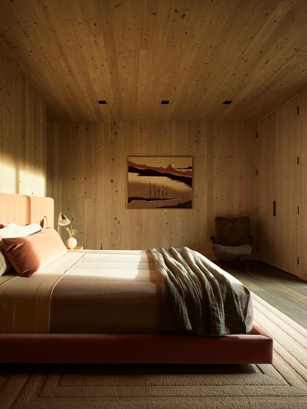 A warm, tranquil bedroom clad almost entirely in natural timber boards, including the walls and ceiling. The room contains a low upholstered bed with pale peach bedding, a textured dark throw, a small bedside lamp and plant, a vintage-style armchair, built-in timber storage, a framed landscape artwork, and a large neutral textured rug. Strong sunlight creates dramatic bands of light across the bed and floor.