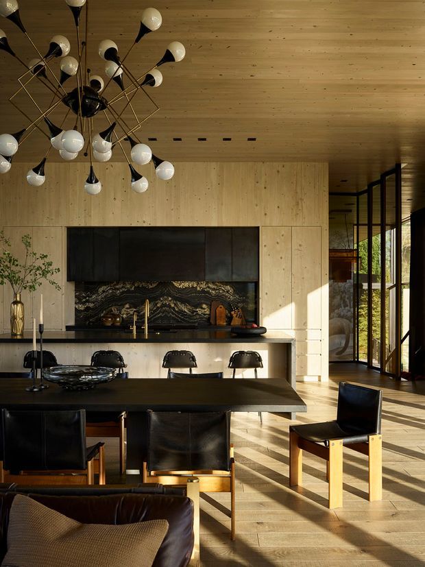 A warm, sophisticated open-plan kitchen and dining area with light timber wall and ceiling panelling, dark cabinetry, a dramatic veined stone backsplash, and a long dark dining table with black leather chairs. A sculptural globe chandelier hangs above, while full-height glazed doors on the right bring in strong sunlight and views of greenery. A living seating area is partially visible in the foreground.