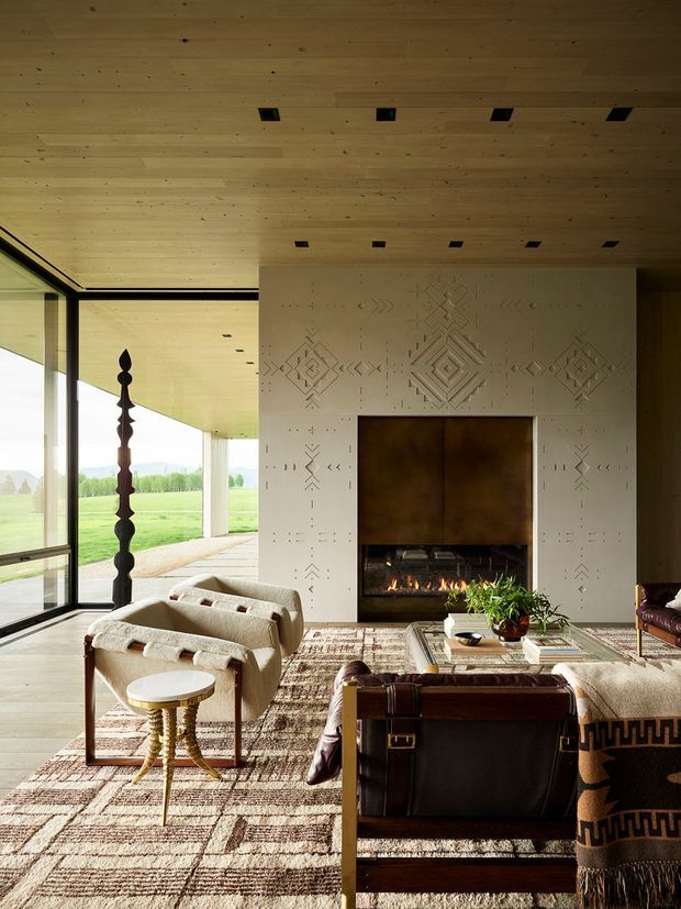 A warm, refined living room centred on a large fireplace with a patterned plaster or concrete feature wall. The room has a timber-lined ceiling with recessed downlights, expansive black-framed glazing opening to a rural landscape, two upholstered lounge chairs, a leather chair, a low coffee table, and a large patterned rug. A sculptural dark timber post and woven textiles add distinctive natural detailing.