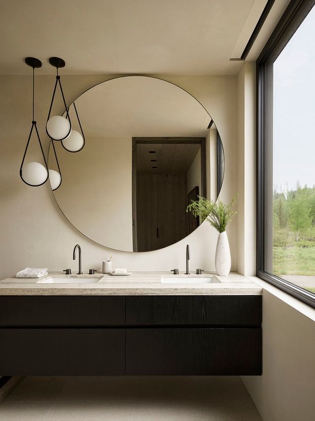 A refined contemporary bathroom vanity with a long double-sink counter, dark wood floating cabinetry, pale stone benchtop, warm beige walls, and a large round mirror. Sculptural black pendant lights hang above the vanity, while a full-height corner window brings in natural light and views of greenery.
