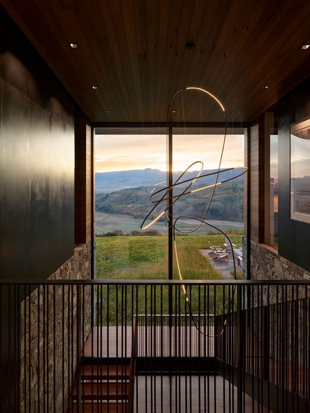 A dramatic interior view of a contemporary multi-level circulation space overlooking a broad rural valley. Dark timber ceiling boards, black metal wall panels, natural stone-clad walls, expansive floor-to-ceiling glazing, and slender black stair balustrades frame a sculptural suspended loop light. A stair descends to a lower level, while outdoor seating and landscaped grounds are visible beyond the glazing. The palette is warm, dark, and natural, with a strong indoor-outdoor connection.