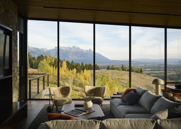A refined living room with expansive floor-to-ceiling glazing overlooking a dramatic mountain and valley landscape. The room features a large sectional sofa, sculptural lounge chairs, a low coffee table, stone wall detailing, warm timber ceiling lining, dark window frames, and understated contemporary furnishings. The composition feels calm, warm, and strongly connected to the surrounding landscape.