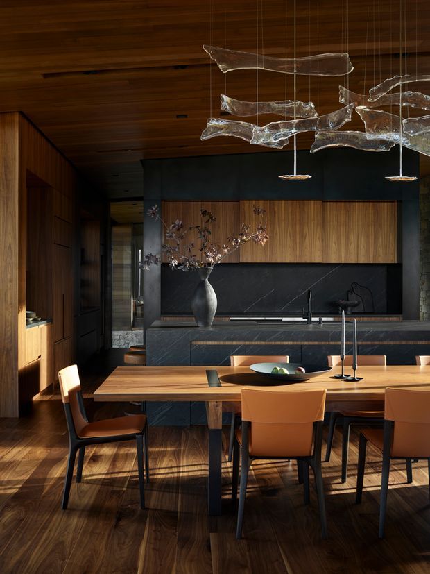 A sophisticated open-plan kitchen and dining area with a dark, richly textured palette. The room features a long timber dining table with leather dining chairs, dark stone kitchen island and splashback, warm timber cabinetry and wall panelling, and a dramatic sculptural glass pendant installation suspended above the dining zone. Timber flooring and ceiling create a cohesive, warm interior, while integrated dark cabinetry and minimal detailing give the space a refined contemporary character.