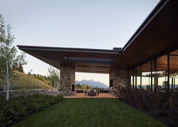 A contemporary mountain residence with broad cantilevered flat roof planes, exposed dark timber soffits, stone-clad structural columns, and extensive floor-to-ceiling glazing. The covered outdoor terrace frames a distant mountain view and connects to a landscaped lawn and garden.