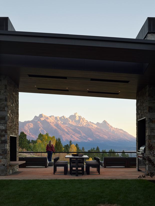 A dramatic covered outdoor entertaining terrace framed by substantial dark stone-clad walls and a broad flat roof canopy. The timber-lined ceiling includes recessed lighting and concealed linear openings. Built-in and freestanding dark outdoor lounge furniture surrounds a dining table, with a built-in barbecue visible at the right. The terrace overlooks a lawn, trees, and a prominent rugged mountain range in warm evening light.