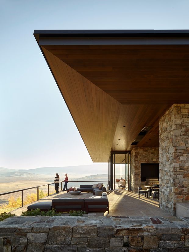 A spacious covered outdoor terrace attached to a modern stone-clad home, overlooking a broad rural valley. The terrace features a dramatic cantilevered timber-lined ceiling and soffit, dark metal roof edging, full-height glazing, stone walls and paving, a large sectional lounge, dining furniture, and a slim black balustrade. The setting is bright and expansive, with warm natural materials and strong indoor-outdoor connection.