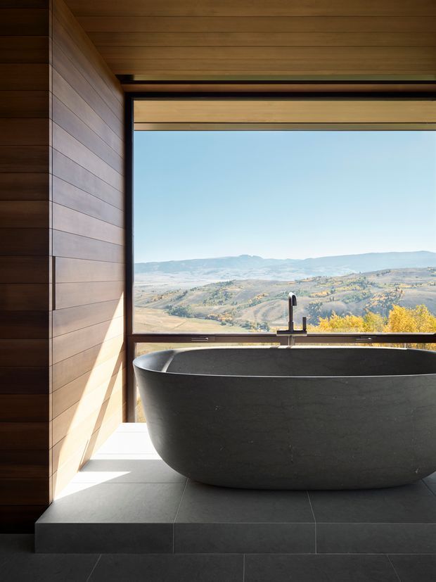 A dark freestanding bathtub is positioned on a raised tiled platform beside a large black-framed picture window, overlooking a broad rural mountain landscape. The bathroom has warm horizontal timber wall and ceiling lining, dark floor tiles, and a minimalist black tap. Strong natural light contrasts with the charcoal bath and finishes.