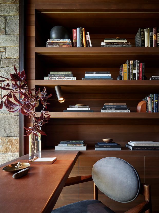 A richly detailed study or home office with full-height dark timber built-in shelving and cabinetry, filled with books and a few sculptural objects. A warm timber desk occupies the foreground beside a rounded upholstered armchair, while a small integrated reading light illuminates the shelves. A stone wall and tall window edge appear at left, with burgundy foliage in a glass vase adding colour. The space has a refined, warm, library-like atmosphere.