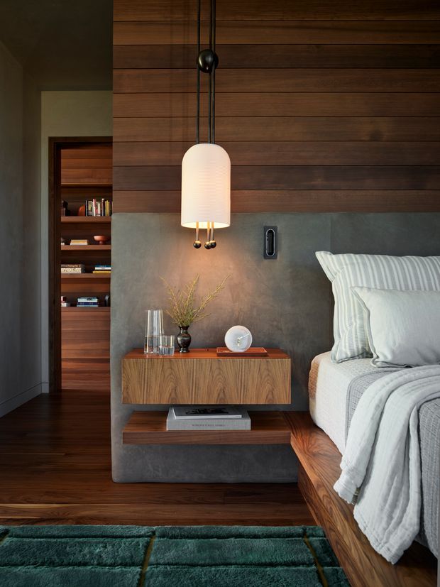 A moody, refined bedroom interior featuring a wood-panelled feature wall, concrete-look upholstered or rendered headboard, integrated timber bedside storage, and a sculptural pendant lamp. The bed has layered white and grey striped bedding, with a deep green rug in the foreground. A recessed timber bookshelf is visible through an opening beside the bed.