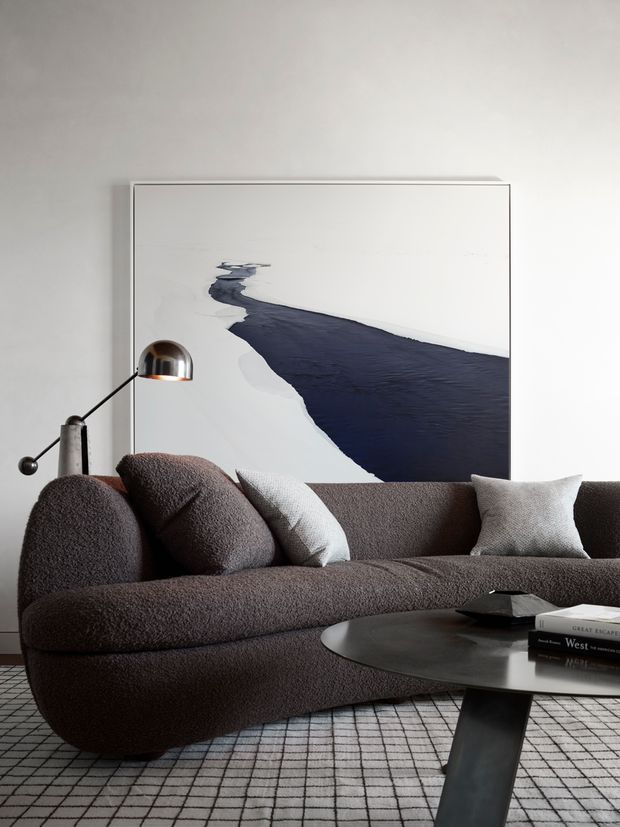 A refined contemporary living room vignette featuring a deep brown textured curved sofa with coordinating neutral cushions, a black oval coffee table, a black-and-white grid rug, and a slim arched floor lamp. A large abstract monochrome artwork provides a dramatic focal point against the pale wall.