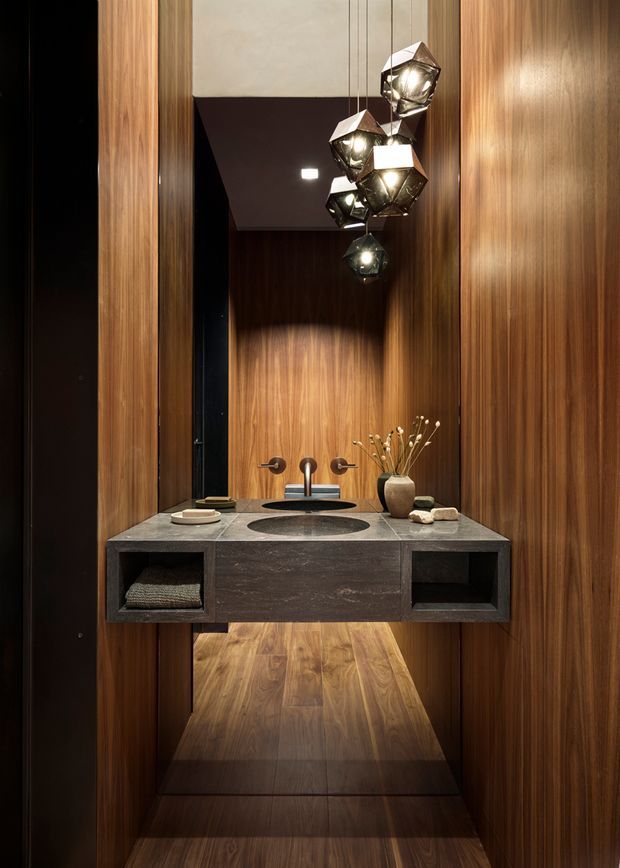 A dramatic, compact bathroom vanity set within a richly timber-lined alcove. The floating dark stone vanity has two round vessel basins, wall-mounted bronze-toned taps, open shelving, and a large mirror. Faceted pendant lights hang in a cluster above, creating warm highlights against the deep wood surfaces. A timber-floored space is visible beyond the vanity.