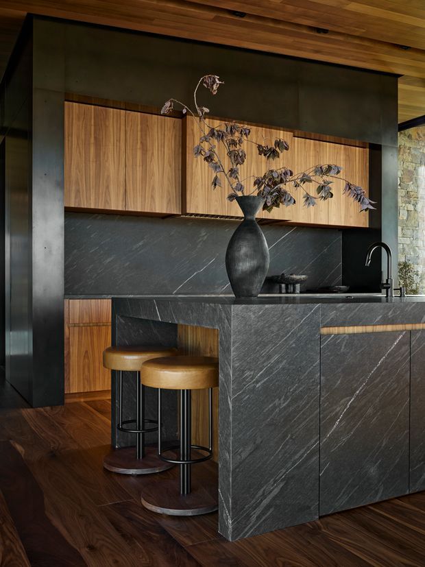 A moody, refined contemporary kitchen with a large dark stone island, integrated black sink and tap, warm timber cabinetry, dark timber flooring, and black structural framing. Two cognac leather bar stools sit at the island, while a sculptural dark vase provides a central decorative accent. A rough stone wall is visible at the right edge, and timber boards line the ceiling.