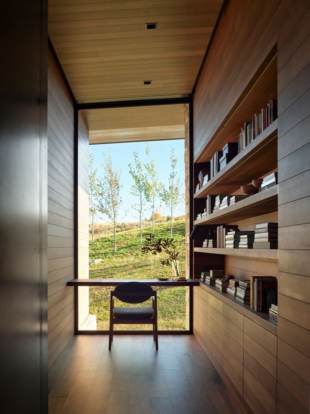 A narrow, warm-toned interior passage or study nook with full-height timber built-in bookshelves along one wall, a slim desk spanning the glazed end, and a single dark chair. Horizontal timber wall and ceiling panelling, dark metal framing, and a large window create a quiet connection to the landscaped outdoors. The space is minimalist and refined, with strong natural light and extensive built-in joinery.