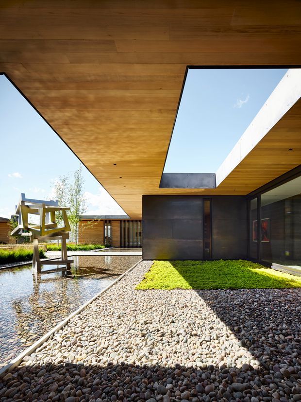 A contemporary courtyard viewed from beneath a broad cantilevered timber-lined roof. The courtyard features pebble landscaping, a shallow reflective water feature, clipped lawn, dark metal-clad walls, extensive glazing, and a sculptural timber installation. Strong geometric roof openings frame the sky and create a dramatic interplay of sunlight and shadow.