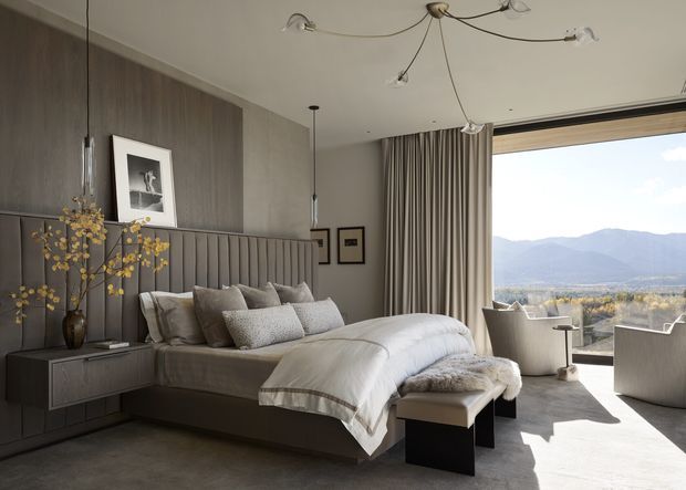 A refined contemporary bedroom with a large upholstered bed, layered neutral bedding, integrated bedside cabinetry, and a vertically panelled grey feature wall. Full-height curtains frame expansive glazing with a mountain landscape beyond. A sculptural multi-arm ceiling light, upholstered bench, lounge chairs, framed artwork, and a decorative vase complete the calm, luxurious setting.