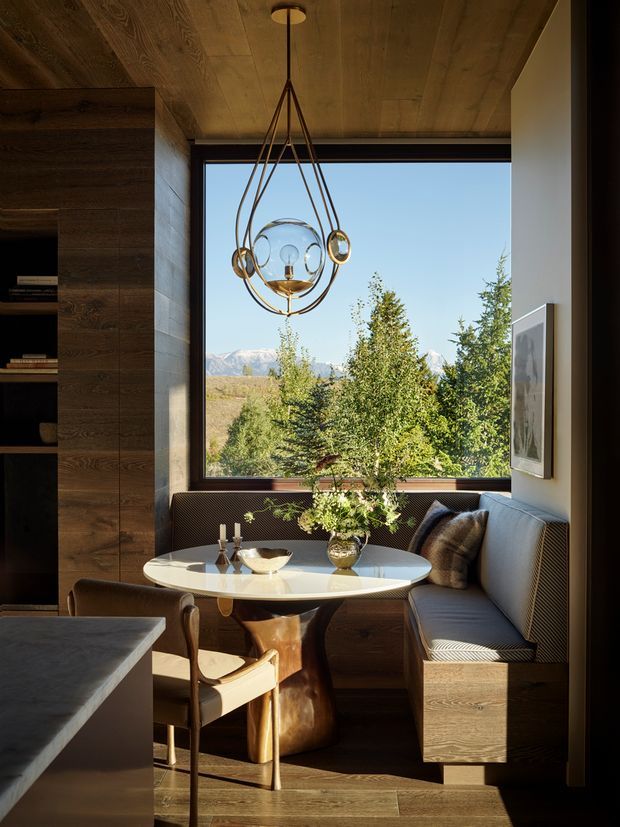 A warm, refined dining nook with a built-in upholstered banquette, round glossy table, sculptural pedestal base, and leather dining chair. Dark timber cabinetry and a timber-lined ceiling frame a large picture window overlooking trees and distant mountains. A distinctive sculptural pendant hangs above the table, while sunlight creates strong natural contrast across the richly toned interior.