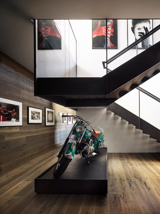 A dramatic contemporary interior featuring a sculptural black steel staircase with exposed timber treads, overlooking a gallery-like hallway. Dark timber wall panelling and matching timber flooring contrast with white walls and expansive glazed stairwell panels. Framed photographic artwork lines the walls, while a vintage green motorcycle is displayed on a raised black plinth in the centre of the space.