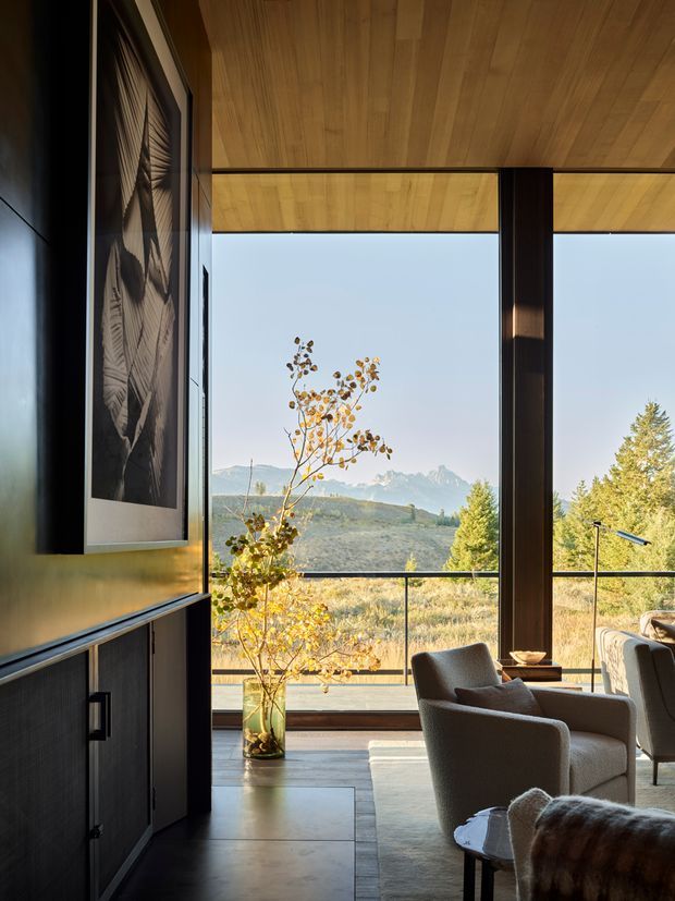 A refined living room with floor-to-ceiling glazing overlooking a dry, mountainous landscape. The space features a warm timber-lined ceiling, dark structural columns and built-in cabinetry, a large monochrome artwork, contemporary upholstered armchairs, a low coffee table, and a glass balustrade along the expansive window wall. Natural daylight and earthy materials create a calm, sophisticated interior.