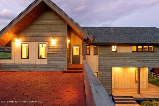 Exterior view of a multi-level timber-clad building at dusk, featuring a prominent steep gable roof, warm illuminated windows and entry doors, exposed timber soffits, and a broad red hexagonal-paver terrace or walkway in the foreground. A lower-level opening and deck are visible to the right, with mountainous terrain faintly visible in the background.