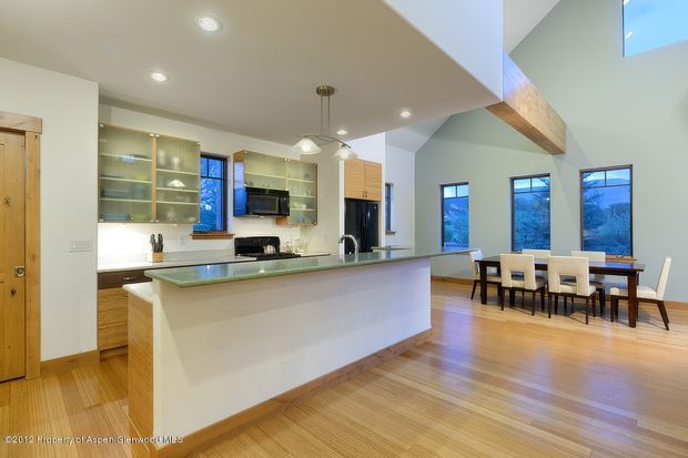 Open-plan kitchen and dining area with a large central island, pale cabinetry, glass-front display shelving, stainless-steel appliances, and a timber floor. The dining space features a rectangular timber table and light upholstered chairs. High angled ceilings, exposed timber structural beams, and expansive windows create a bright, airy interior.