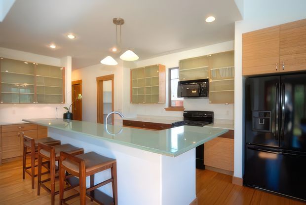 Bright, open kitchen with a large pale glass-topped island and integrated sink, timber cabinetry, glass-front wall cupboards, black appliances, and timber flooring. The island includes seating for four on wooden bar stools, while pendant lights and recessed ceiling lighting provide illumination. A dining or living area is not visible; the image is focused on the kitchen fit-out.