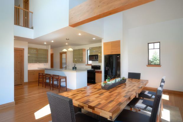 Bright open-plan kitchen and dining area with a large timber dining table in the foreground, a white island with bar stools, extensive built-in cabinetry, timber flooring, and a high ceiling with exposed timber beams. A small upper-level railing and open void are visible at the left, contributing to the spacious interior.