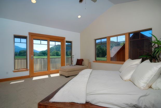 Bright bedroom with a large white bed, beige chaise lounge, warm timber-framed windows and glazed doors, and views toward a green mountainous landscape. The room has a simple, airy layout with light walls, carpeted flooring, and a dark timber bed frame and headboard.