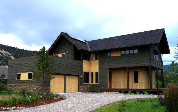 Exterior view of a large contemporary mountain home with dark horizontal wood cladding, contrasting natural timber doors and window shutters, stone base walls, and multiple intersecting pitched gables. The house includes an attached garage and sits beside a gravel driveway with landscaped planting and mountainous surroundings.