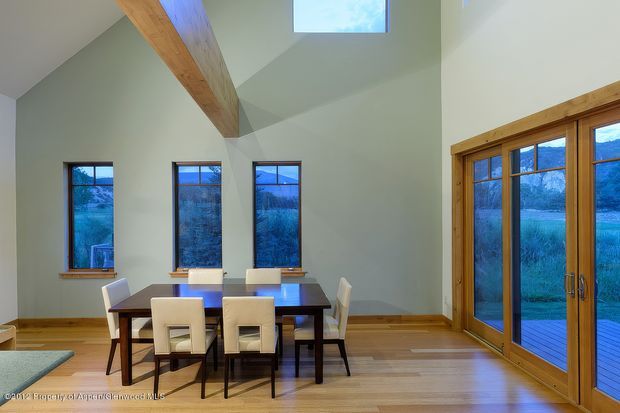 Bright, double-height dining area with a dark rectangular dining table and upholstered chairs, surrounded by tall windows and timber-framed glazed doors opening to a deck. The room has pale green walls, warm timber flooring and exposed timber ceiling beams, with a high clerestory-style skylight/window providing additional daylight. A natural landscape is visible outside.
