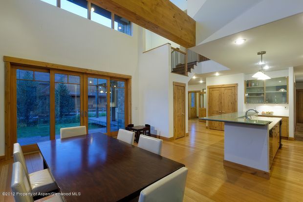 A spacious open-plan interior combining a dining area and kitchen beneath a double-height ceiling. The room features warm timber flooring, exposed timber ceiling beams, extensive timber-framed glazing and doors, a large dark wood dining table, white dining chairs, and a central kitchen island with a light benchtop. A stairway and upper-level landing are visible in the background, along with built-in glass-fronted cabinetry.